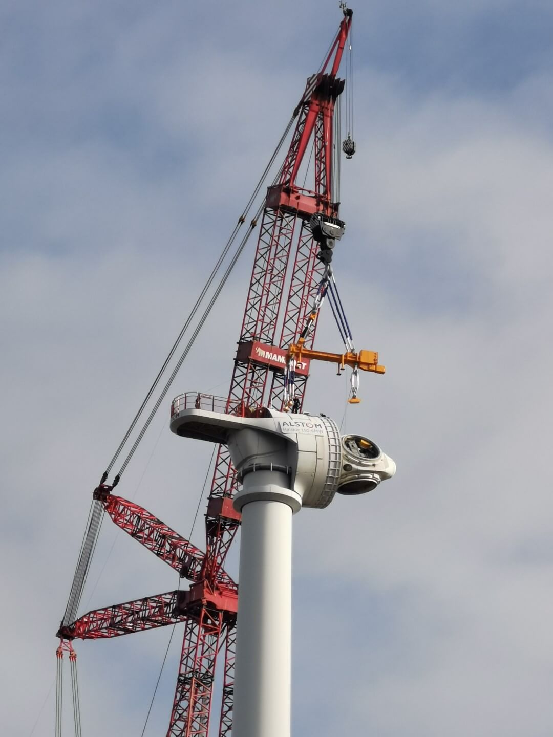 Wind turbine nacelle being lifted and positioned on top of a tower by a large red lattice boom crane during wind farm installation, with rigging equipment and spreader beam visible against cloudy sky.