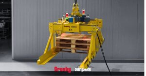 Custom crane pallet lifter by Crosby Airpes — yellow below-the-hook pallet lifting clamp with articulated side arms gripping a wooden pallet, suspended from an overhead crane hook inside an industrial facility.